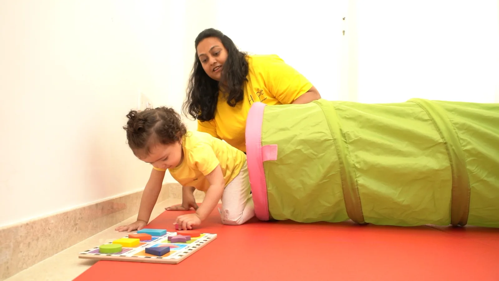 A toddler crawling towards a play tunnel while a therapist cheers her on