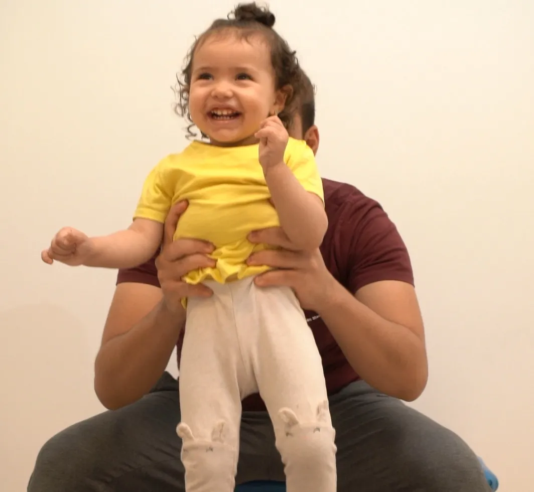 A smiling toddler is held up by her therapist during a movement session