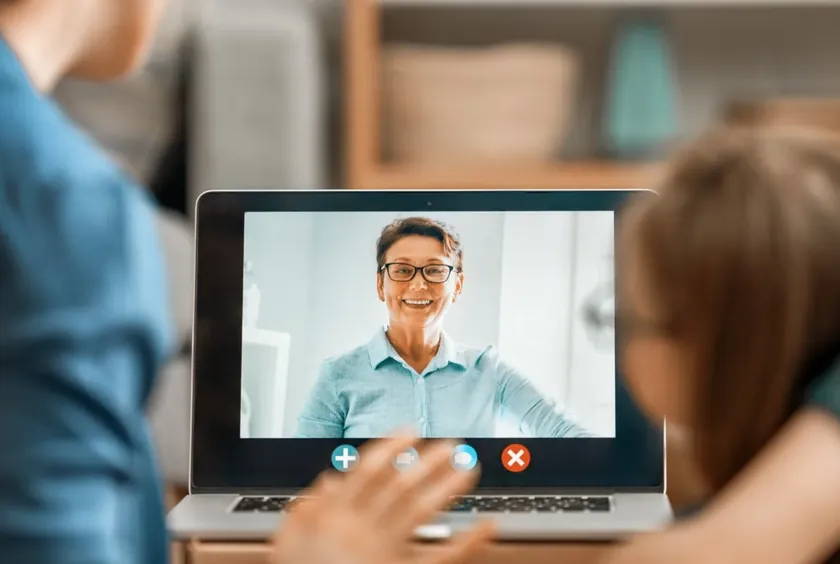 A child and parent wave to a smiling therapist on a laptop video call