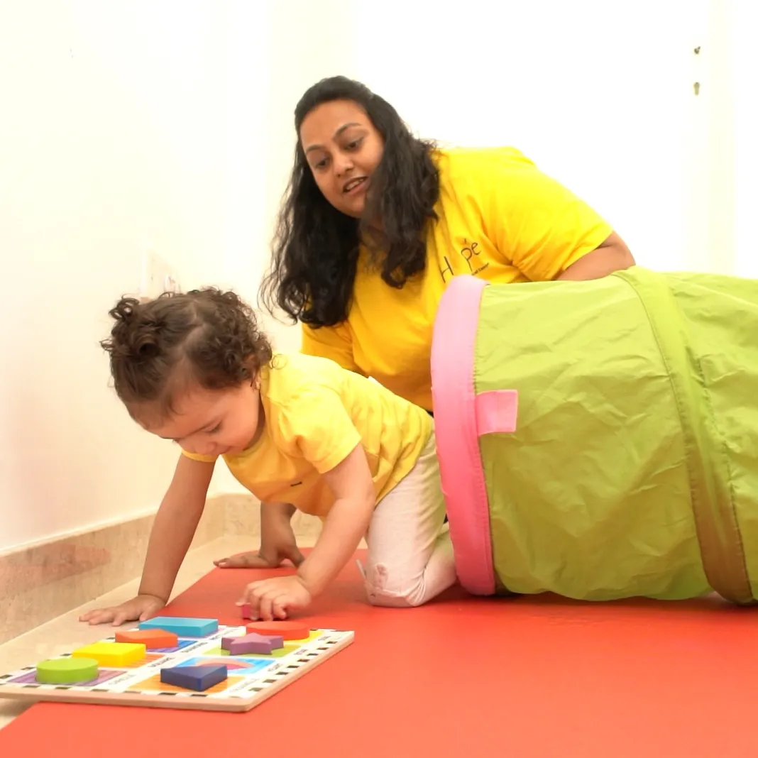 A toddler plays with a shape puzzle as her therapist joins in from the play tunnel