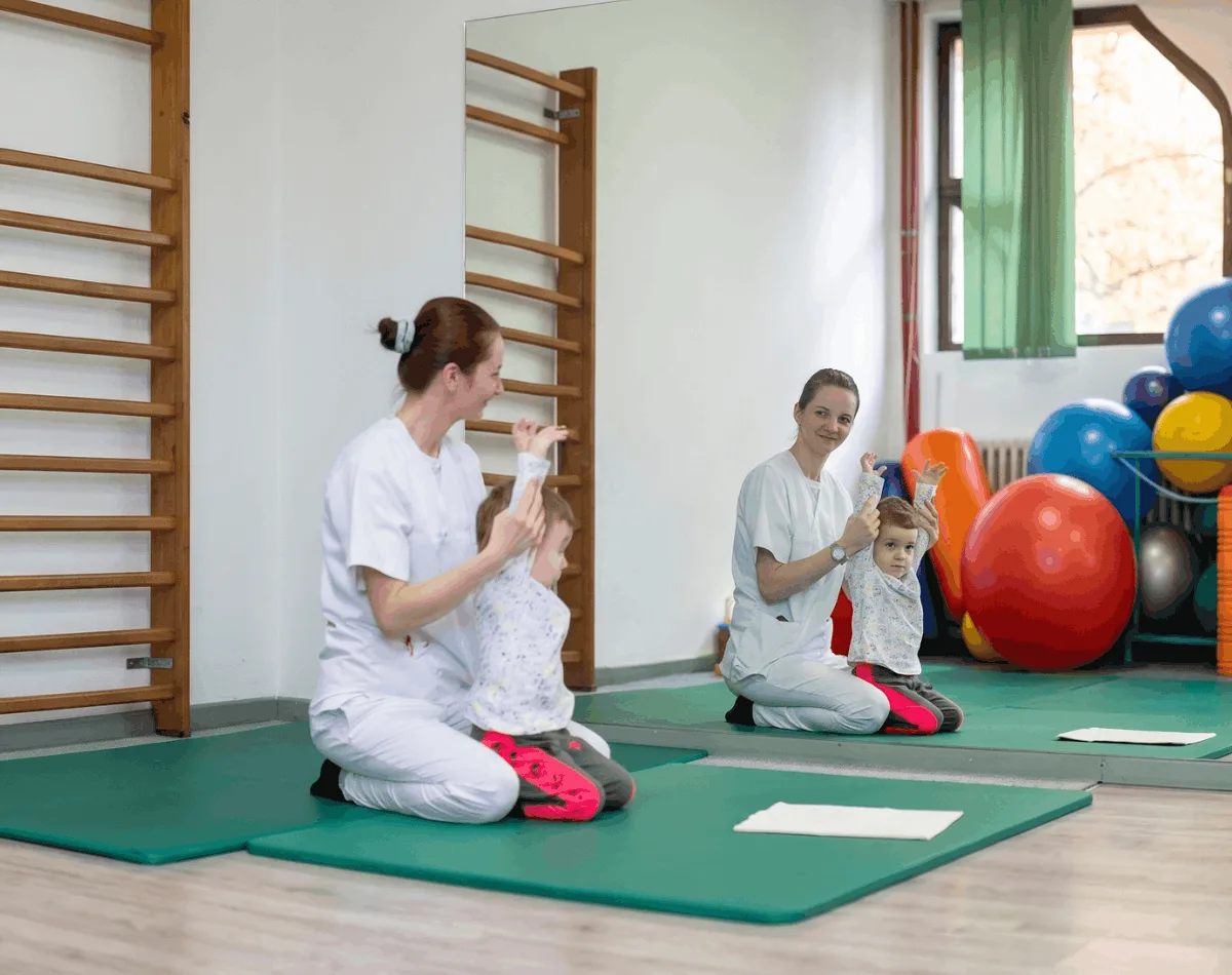 A therapist helps a toddler stretch both arms overhead on an exercise mat in front of a mirror